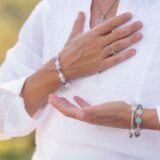Close-up of a hand wearing pastel beaded bracelet and silver rings on a white embroidered blouse, outdoors in soft light.